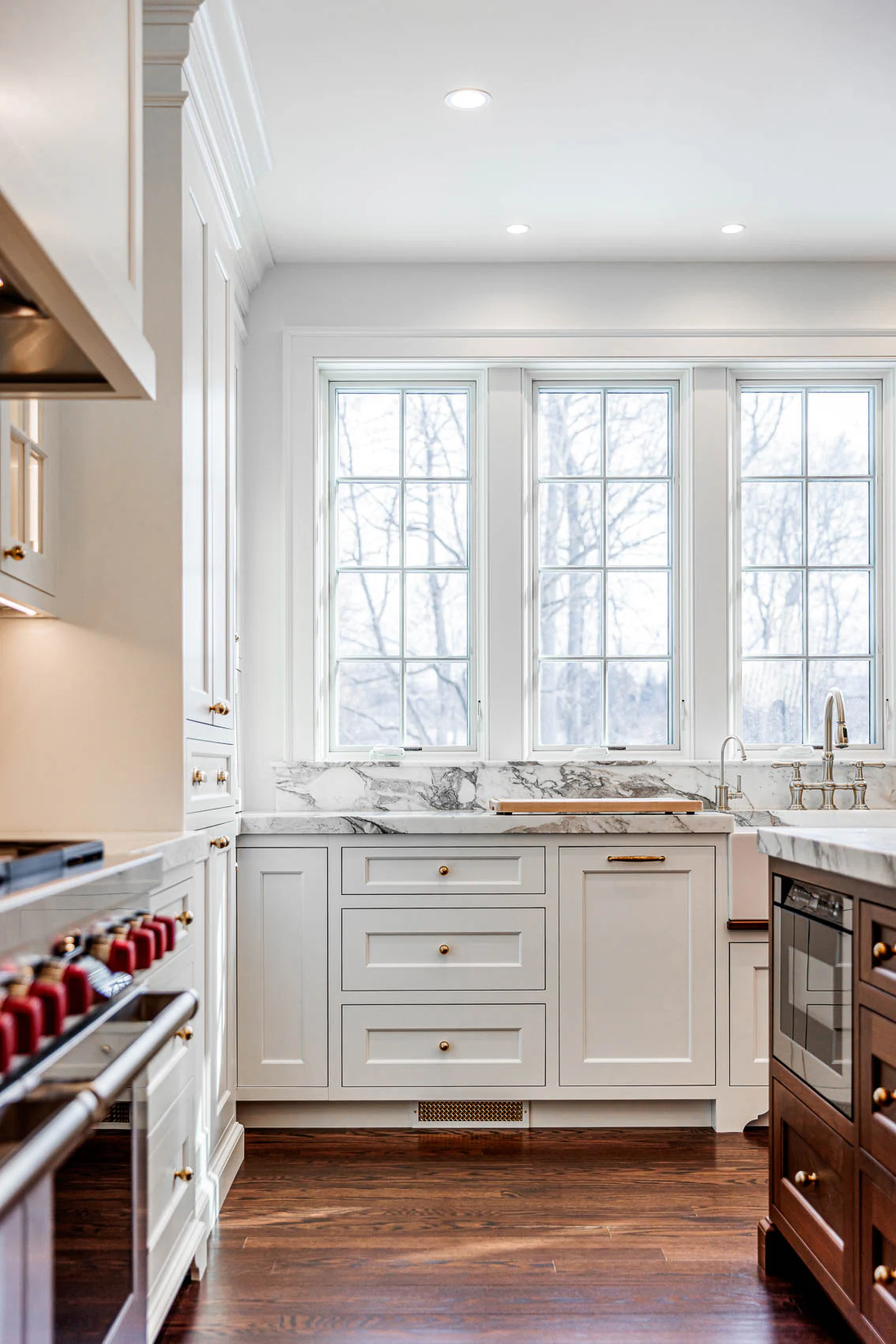 Bright white kitchen with marble countertops, white cabinets, brass hardware, and a double window over the sink showing a snowy outdoor scene.